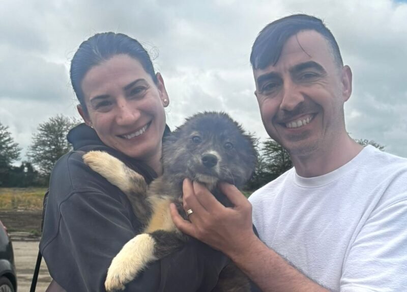Smiling couple holding a fluffy puppy outdoors against a cloudy skyp