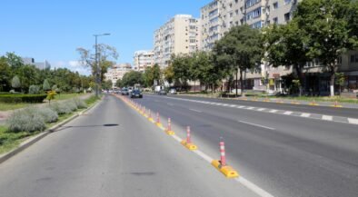 Wide city street with a central row of orange-and-white traffic posts and a sidewalk on the left, tall apartment buildings on the right.