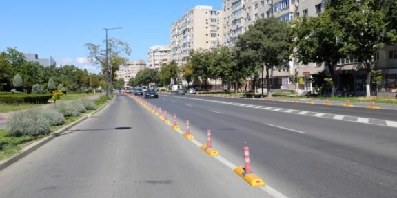 Wide city street with a central row of orange-and-white traffic posts and a sidewalk on the left, tall apartment buildings on the right.