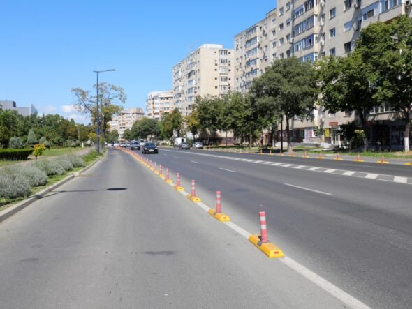 Wide city street with a central row of orange-and-white traffic posts and a sidewalk on the left, tall apartment buildings on the right.