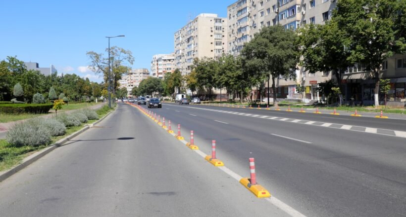 Wide city street with a central row of orange-and-white traffic posts and a sidewalk on the left, tall apartment buildings on the right.