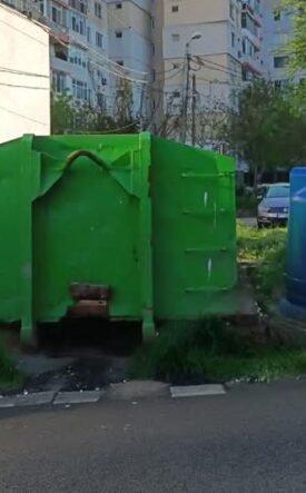 Row of colorful municipal recycling bins and a large green dumpster in a residential area with apartment buildings in the background.