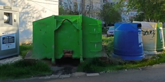 Row of colorful municipal recycling bins and a large green dumpster in a residential area with apartment buildings in the background.