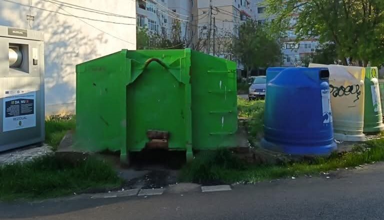 Row of colorful municipal recycling bins and a large green dumpster in a residential area with apartment buildings in the background.