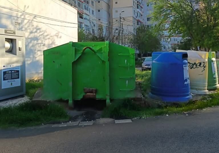 Row of colorful municipal recycling bins and a large green dumpster in a residential area with apartment buildings in the background.