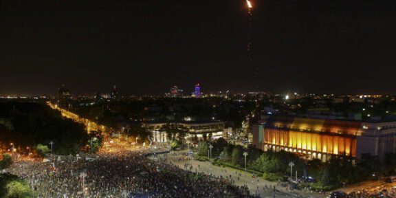 Nighttime aerial view of a crowded city square with a brightly lit building to the right and a tall flame rising into the sky from a rocket or flare above the city.