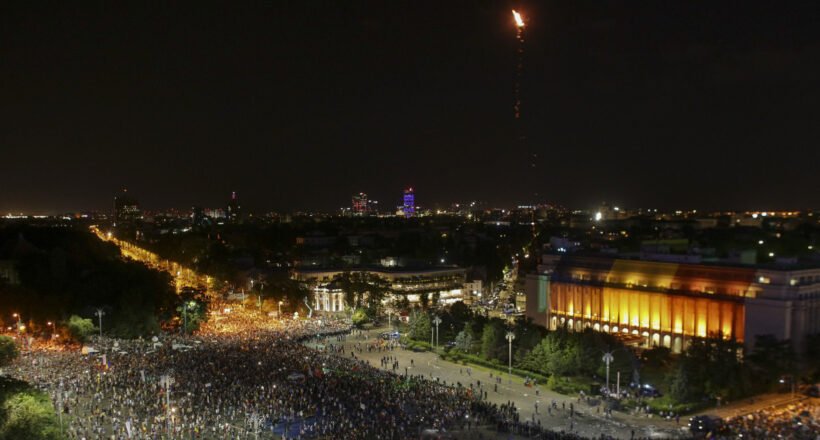Nighttime aerial view of a crowded city square with a brightly lit building to the right and a tall flame rising into the sky from a rocket or flare above the city.
