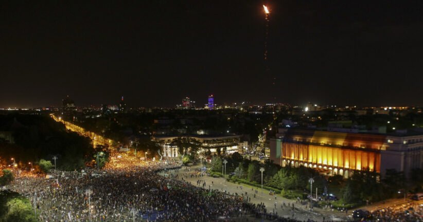 Nighttime aerial view of a crowded city square with a brightly lit building to the right and a tall flame rising into the sky from a rocket or flare above the city.