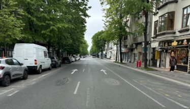 Urban street view with parked cars on both sides, green trees, and a shopfront on the right sidewalk aisle ahead in the distance traffic arrows on the road calm and overcast sky behind trees and buildings.