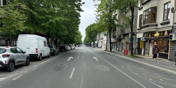 Urban street view with parked cars on both sides, green trees, and a shopfront on the right sidewalk aisle ahead in the distance traffic arrows on the road calm and overcast sky behind trees and buildings.
