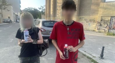 Two young people standing on a street, each holding a bottle/drink can, with a graffiti-covered wall behind them and a parked car nearby.