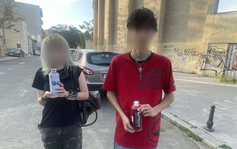 Two young people standing on a street, each holding a bottle/drink can, with a graffiti-covered wall behind them and a parked car nearby.