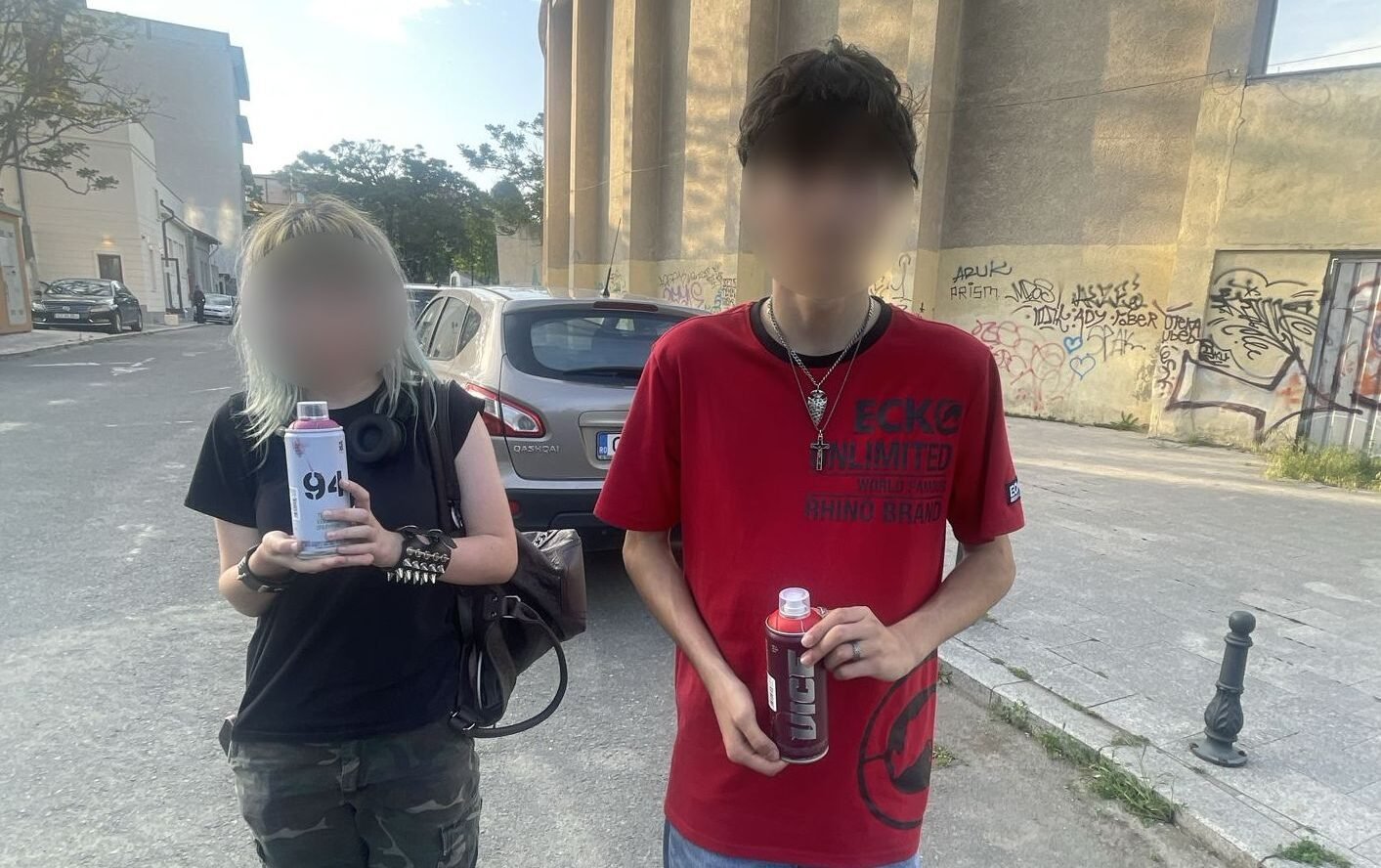 Two young people standing on a street, each holding a bottle/drink can, with a graffiti-covered wall behind them and a parked car nearby.