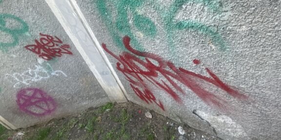 Graffiti-covered concrete wall with red and green spray paint tags near a corner, ground with weeds and debris shows at the base.