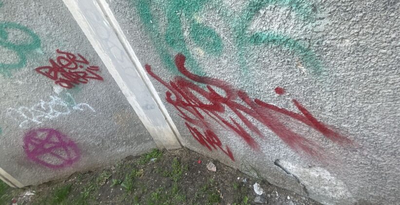 Graffiti-covered concrete wall with red and green spray paint tags near a corner, ground with weeds and debris shows at the base.