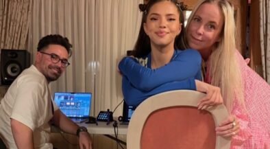 Three people pose for a photo indoors: a man with DJ equipment sits at a table with laptops, while a young woman in blue holds a large circular prop and an older woman in pink stands beside her.