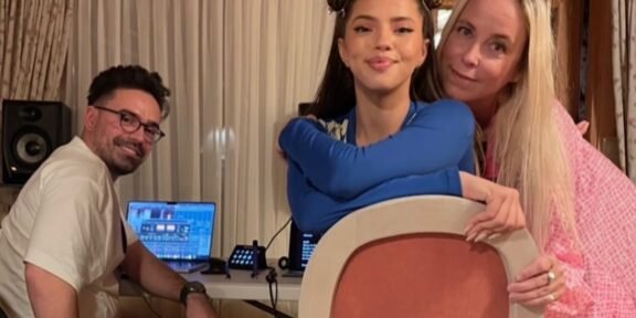 Three people pose for a photo indoors: a man with DJ equipment sits at a table with laptops, while a young woman in blue holds a large circular prop and an older woman in pink stands beside her.