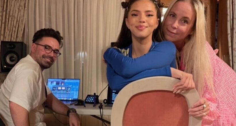 Three people pose for a photo indoors: a man with DJ equipment sits at a table with laptops, while a young woman in blue holds a large circular prop and an older woman in pink stands beside her.