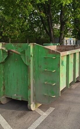 Row of green dumpsters parked along a street beside trees on a sunny day