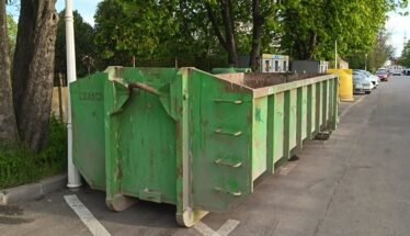 Row of green dumpsters parked along a street beside trees on a sunny day
