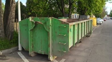 Row of green dumpsters parked along a street beside trees on a sunny day