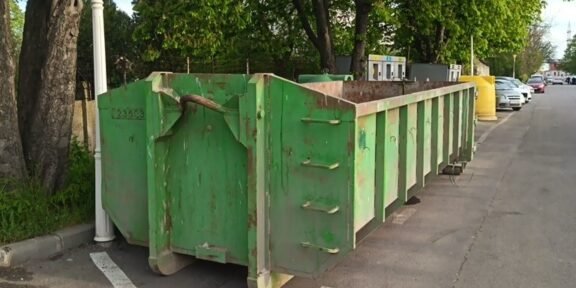 Row of green dumpsters parked along a street beside trees on a sunny day