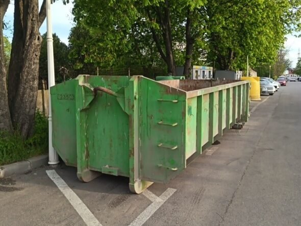 Row of green dumpsters parked along a street beside trees on a sunny day