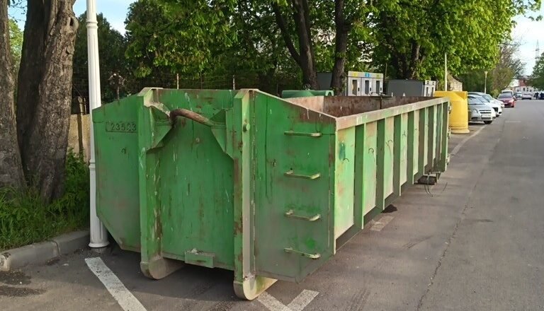 Row of green dumpsters parked along a street beside trees on a sunny day
