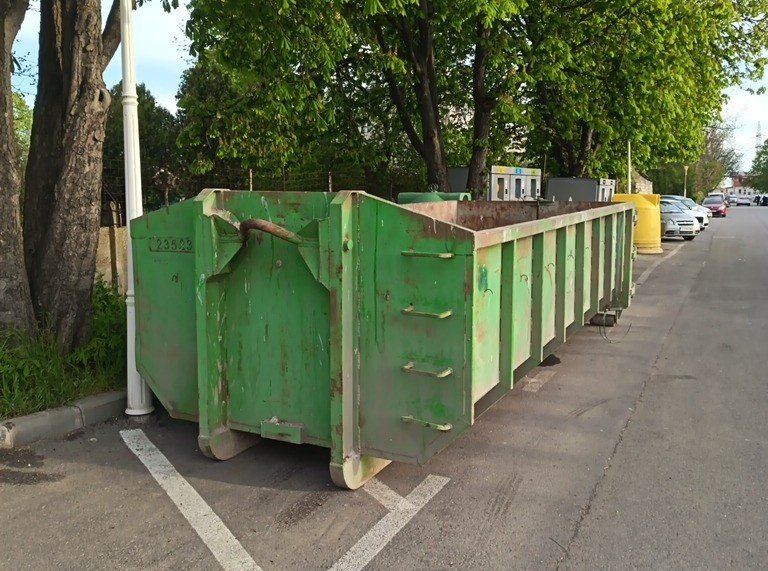 Row of green dumpsters parked along a street beside trees on a sunny day