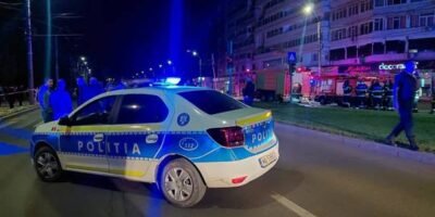 Blue-and-white Romanian police car with flashing blue lights on a nighttime city street, police and bystanders in the background.