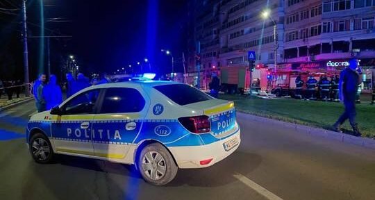 Romanian police car with blue lights on a nighttime city street; bystanders and a lit storefront in the background.