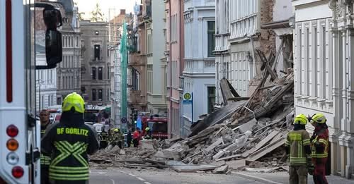 Firefighters in yellow helmets and reflective jackets assess a street blocked by debris from a collapsed building.