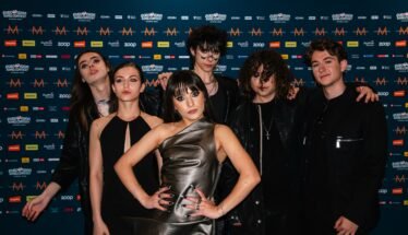 Group of seven young adults posing together in front of a blue, logo-filled backdrop at a media event.