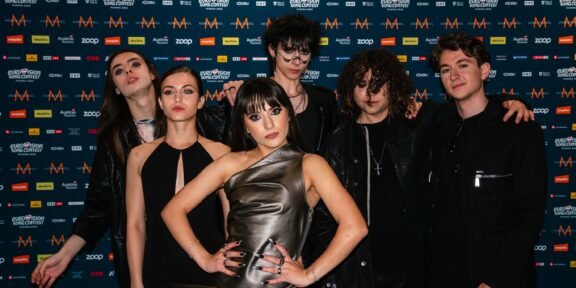 Group of seven young adults posing together in front of a blue, logo-filled backdrop at a media event.