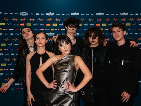 Group of seven young adults posing together in front of a blue, logo-filled backdrop at a media event.