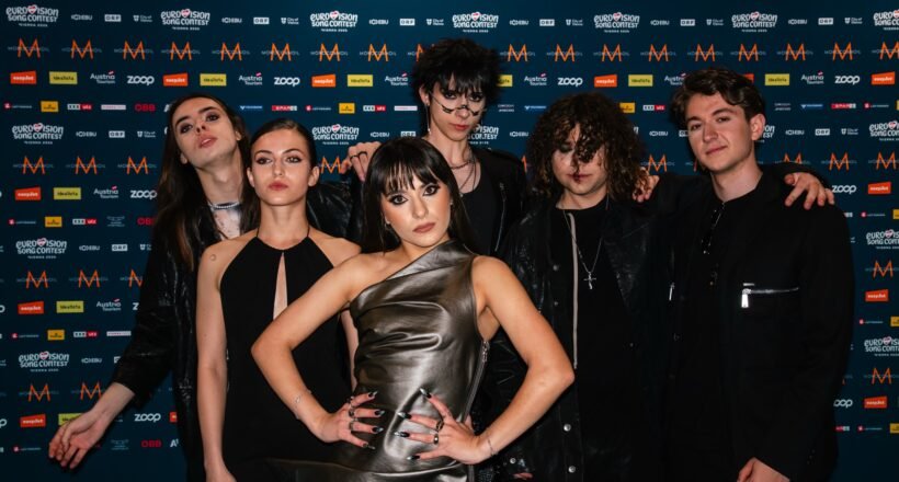 Group of seven young adults posing together in front of a blue, logo-filled backdrop at a media event.