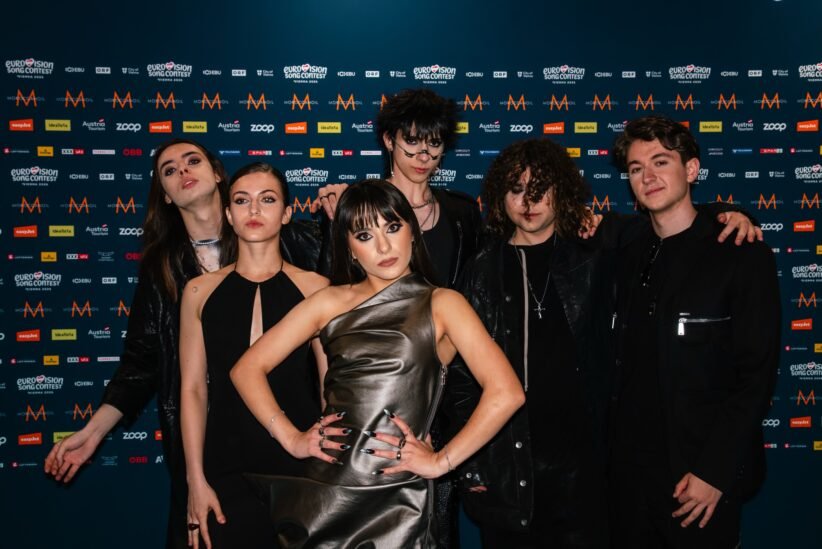 Group of seven young adults posing together in front of a blue, logo-filled backdrop at a media event.