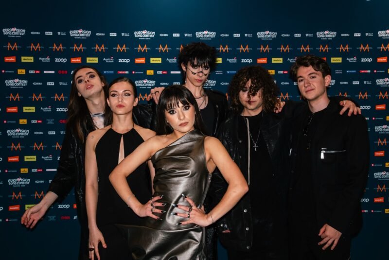 Group of seven young adults posing together in front of a blue, logo-filled backdrop at a media event.
