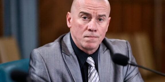 Bald man in a gray suit and striped tie speaks into a microphone at a formal hearing.