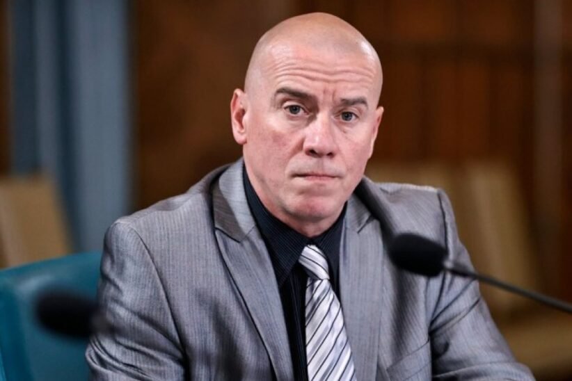 Bald man in a gray suit and striped tie speaks into a microphone at a formal hearing.