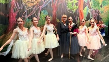Group of young ballet dancers in pastel tutus posing with two adults on a stage with a forest backdrop of trees and greenery.
