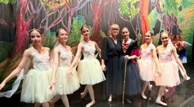 Group of young ballet dancers in pastel tutus posing with two adults on a stage with a forest backdrop of trees and greenery.