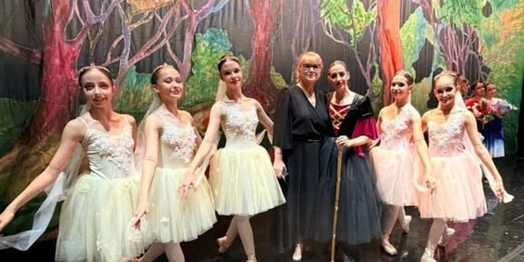 Group of young ballet dancers in pastel tutus posing with two adults on a stage with a forest backdrop of trees and greenery.