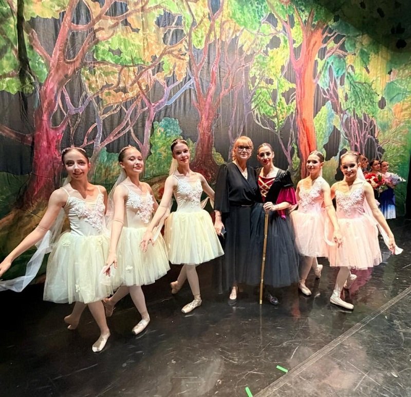Group of young ballet dancers in pastel tutus posing with two adults on a stage with a forest backdrop of trees and greenery.