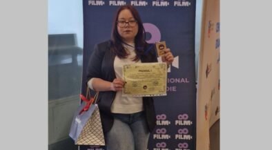 Woman with glasses in a blazer and white top holds a certificate and a trophy in front of a blue film festival banner, with a gift bag at her side.