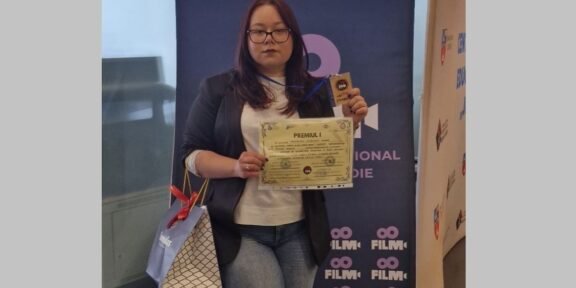 Woman with glasses in a blazer and white top holds a certificate and a trophy in front of a blue film festival banner, with a gift bag at her side.