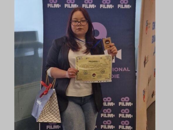 Woman with glasses in a blazer and white top holds a certificate and a trophy in front of a blue film festival banner, with a gift bag at her side.
