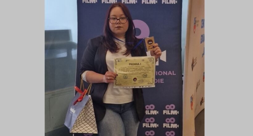 Woman with glasses in a blazer and white top holds a certificate and a trophy in front of a blue film festival banner, with a gift bag at her side.