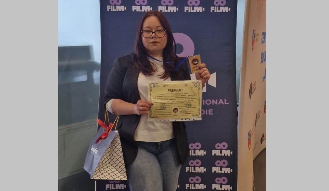 Woman with glasses in a blazer and white top holds a certificate and a trophy in front of a blue film festival banner, with a gift bag at her side.