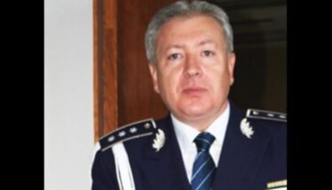Middle-aged man in formal max blue military uniform with epaulets, medals, and striped tie, standing indoors.
